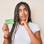 Young african american woman with braids holding birth control pills serious face thinking about question with hand on chin, thoughtful about confusing idea